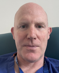 Man looking at the camera, wearing blue scrubs and a lanyard